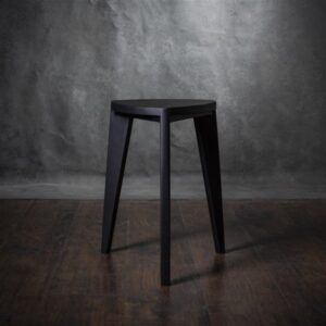 Crna drvena hoklica sa sedalom u obliku Reuleaux trougla i tri blago zakošene noge, fotografisana na tamnom drvenom podu uz sivi betonski zid u pozadini.