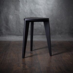 Crna drvena hoklica sa sedalom u obliku Reuleaux trougla i tri blago zakošene noge, fotografisana na tamnom drvenom podu uz sivi betonski zid u pozadini.