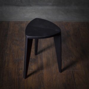 Crna drvena hoklica sa sedalom u obliku Reuleaux trougla i tri blago zakošene noge, fotografisana na tamnom drvenom podu uz sivi betonski zid u pozadini.