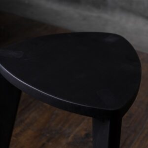 Crna drvena hoklica sa sedalom u obliku Reuleaux trougla i tri blago zakošene noge, fotografisana na tamnom drvenom podu uz sivi betonski zid u pozadini.