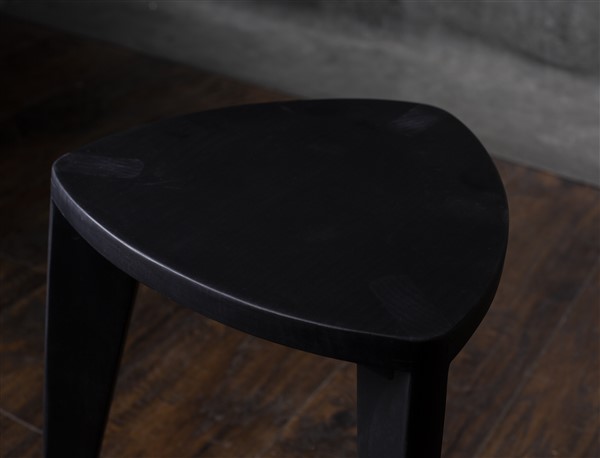 Crna drvena hokllica Crna drvena hoklica sa sedalom u obliku Reuleaux trougla i tri blago zakošene noge, fotografisana na tamnom drvenom podu uz sivi betonski zid u pozadini.