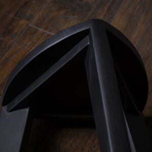 Crna drvena hoklica sa sedalom u obliku Reuleaux trougla i tri blago zakošene noge, fotografisana na tamnom drvenom podu uz sivi betonski zid u pozadini.