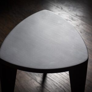 Crna drvena hoklica sa sedalom u obliku Reuleaux trougla i tri blago zakošene noge, fotografisana na tamnom drvenom podu uz sivi betonski zid u pozadini.