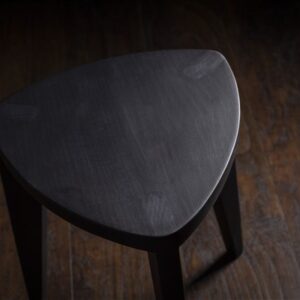 Crna drvena hoklica sa sedalom u obliku Reuleaux trougla i tri blago zakošene noge, fotografisana na tamnom drvenom podu uz sivi betonski zid u pozadini.