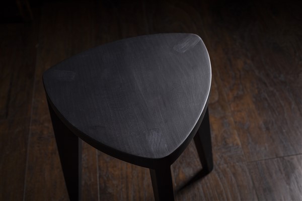 Crna drvena hoklica Crna drvena hoklica sa sedalom u obliku Reuleaux trougla i tri blago zakošene noge, fotografisana na tamnom drvenom podu uz sivi betonski zid u pozadini.