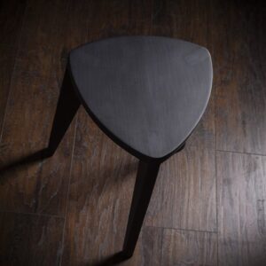 Crna drvena hoklica sa sedalom u obliku Reuleaux trougla i tri blago zakošene noge, fotografisana na tamnom drvenom podu uz sivi betonski zid u pozadini.