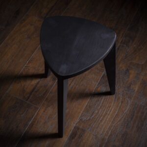 Crna drvena hoklica sa sedalom u obliku Reuleaux trougla i tri blago zakošene noge, fotografisana na tamnom drvenom podu uz sivi betonski zid u pozadini.