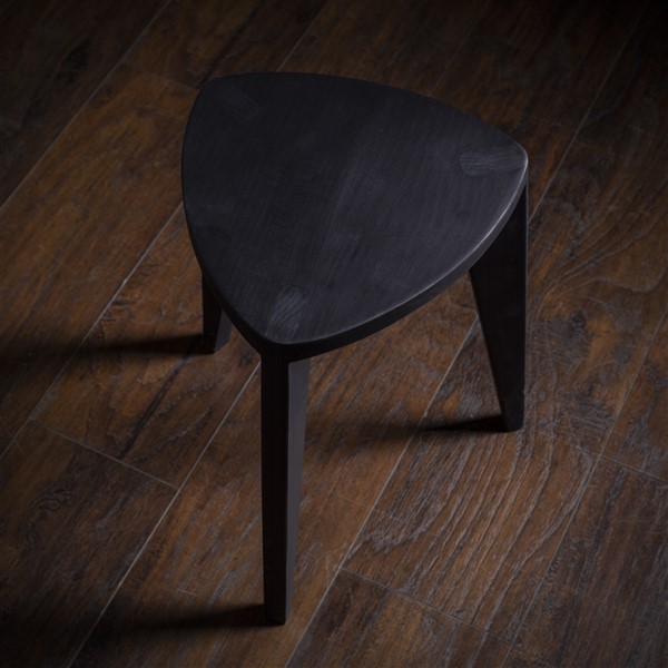 Crna drvena hoklica Crna drvena hoklica sa sedalom u obliku Reuleaux trougla i tri blago zakošene noge, fotografisana na tamnom drvenom podu uz sivi betonski zid u pozadini.