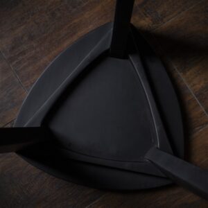 Crna drvena hoklica sa sedalom u obliku Reuleaux trougla i tri blago zakošene noge, fotografisana na tamnom drvenom podu uz sivi betonski zid u pozadini.