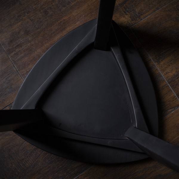 Crna drvena hoklica Crna drvena hoklica sa sedalom u obliku Reuleaux trougla i tri blago zakošene noge, fotografisana na tamnom drvenom podu uz sivi betonski zid u pozadini.